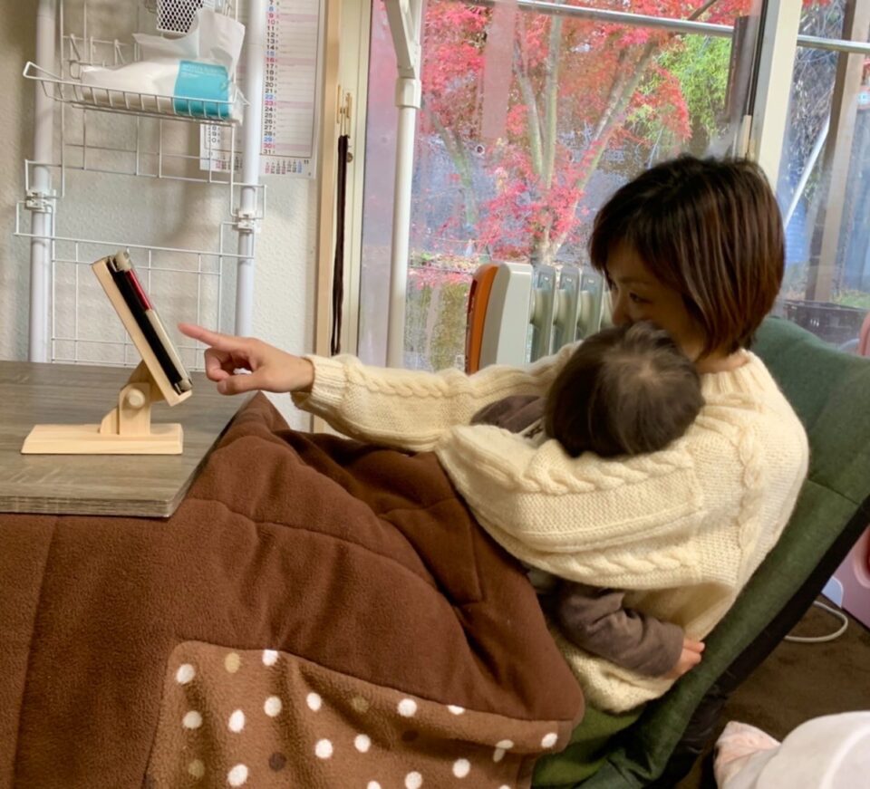 ママが赤ちゃんを抱っこしながらこたつでスマホスタンドのスマホを操作。